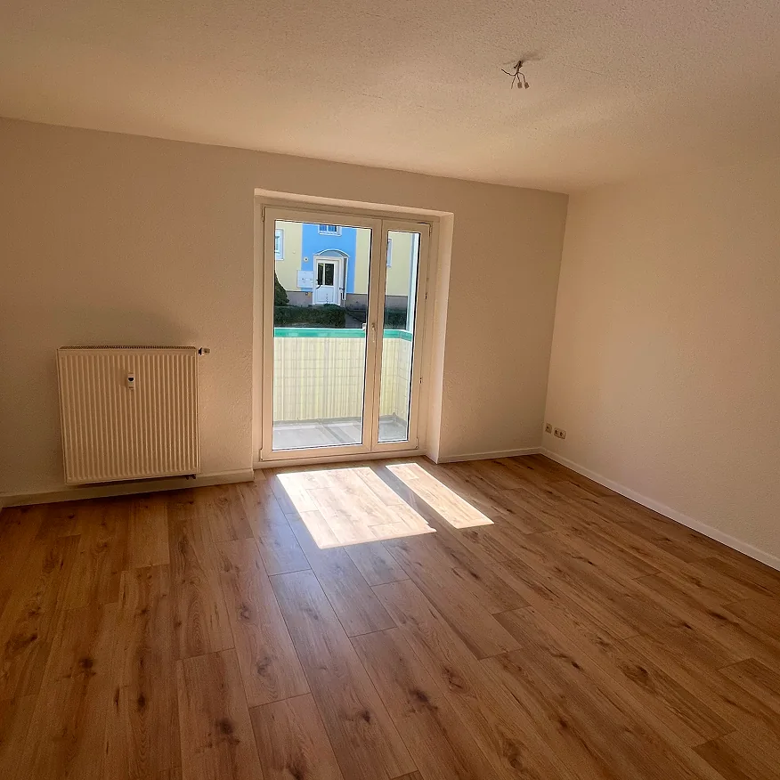 Leerer Raum mit Holzboden, einer Heizung und einer Glastür zu einem kleinen Balkon.